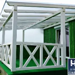 Biała drewniana konstrukcja z zadaszeniem, prawdopodobnie pergola, z balustradą w kształcie krzyży, przylegająca do zielonej ściany budynku, na sztucznej trawie.