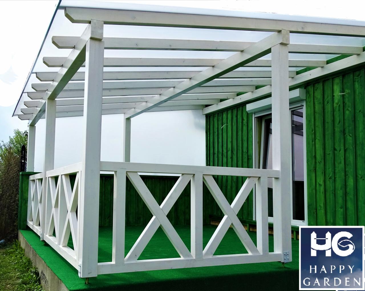Biała drewniana konstrukcja z zadaszeniem, prawdopodobnie pergola, z balustradą w kształcie krzyży, przylegająca do zielonej ściany budynku, na sztucznej trawie.