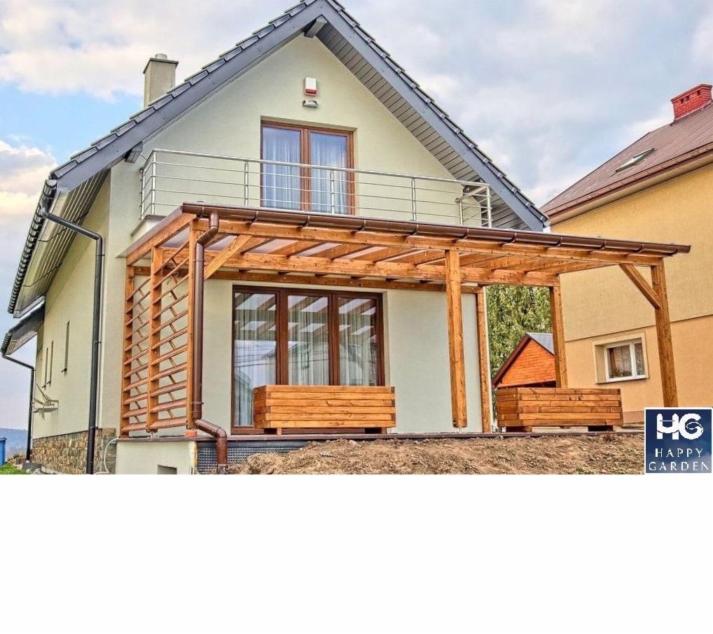 Drewniana pergola z ażurową ścianką boczną, przylegająca do domu z balkonem, z drewnianymi donicami u podstawy, na tle sąsiedniego budynku.