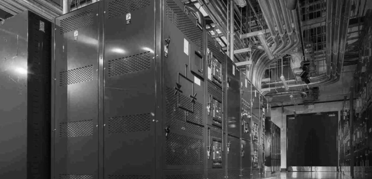 Rząd metalowych szaf elektrycznych z perforowanymi drzwiami w przemysłowym pomieszczeniu, widoczne rury i przewody pod sufitem.