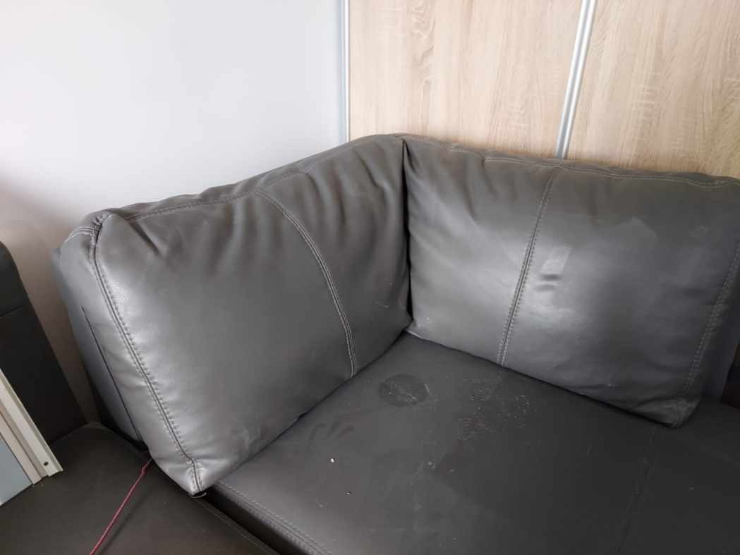 Fragment narożnika z szarej skóry ekologicznej przed renowacją, widoczne plamy i ślady użytkowania na siedzisku, szwy dekoracyjne.