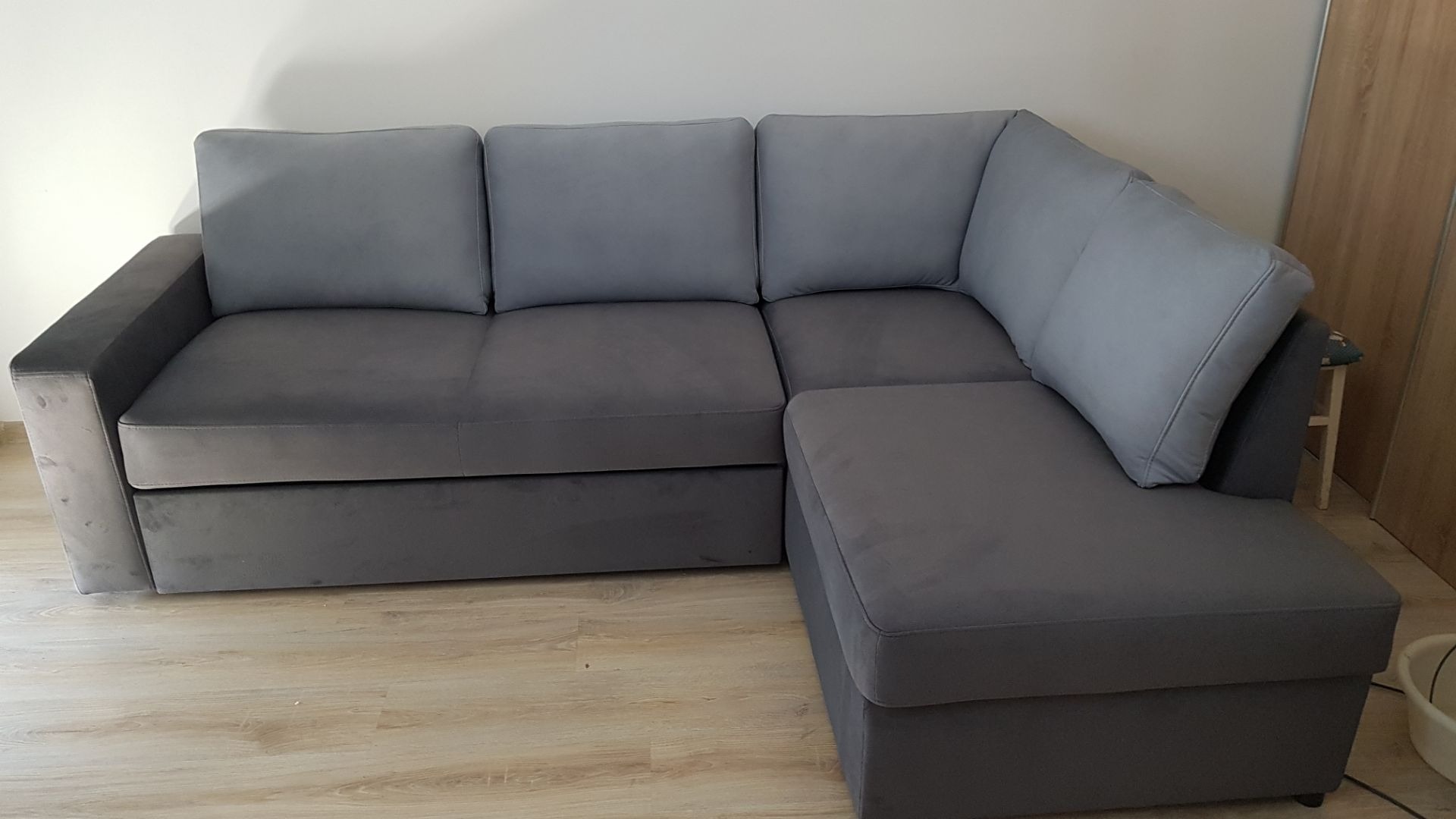 Szary narożnik z trzema poduszkami po renowacji, widoczny na tle jasnej ściany i drewnianej podłogi.