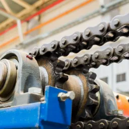 Zbliżenie na przekładnię łańcuchową z dwoma łańcuchami, widoczny mechanizm zębaty i elementy mocujące w odcieniach szarości, niebieskiego i pomarańczowego, w tle konstrukcja hali.