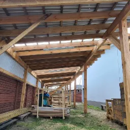 Wnętrze wiaty z drewnianą konstrukcją nośną, częściowo zadaszonej blachą falistą i przezroczystymi płytami, widoczne materiały budowlane i narzędzia, ogrodzenie z cegły betonowej w tle.