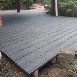 Szara, deszczułkowa powierzchnia tarasu, z widoczną konstrukcją nośną, częściowo obłożona kamieniami, z fragmentem ściany z czerwonej cegły w tle.