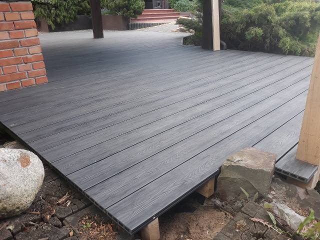 Szara, deszczułkowa powierzchnia tarasu, z widoczną konstrukcją nośną, częściowo obłożona kamieniami, z fragmentem ściany z czerwonej cegły w tle.