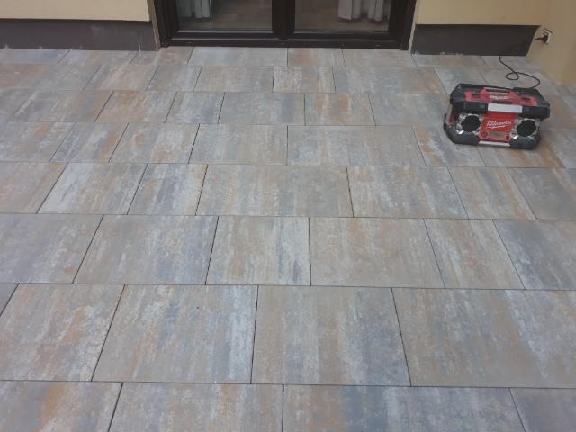 Wyłożony kostką betonową taras przy budynku, widoczny radioodtwarzacz budowlany w rogu.