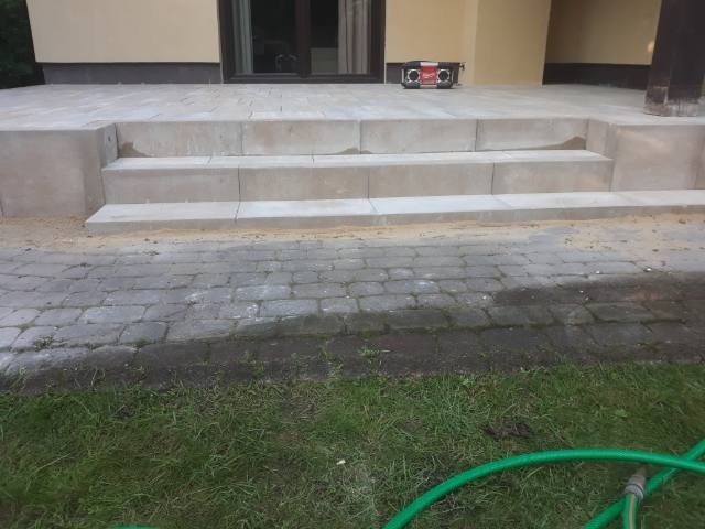 Schody wejściowe z jasnych, betonowych bloków prowadzące do budynku, widoczne kostka brukowa i trawnik z wężem ogrodowym.