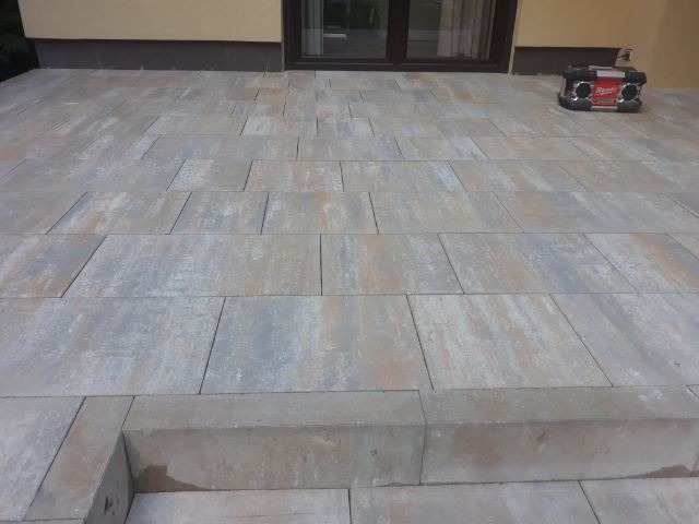 Wykończony taras z szarych płyt betonowych o nieregularnym kształcie, z niskimi schodami wejściowymi i radiem budowlanym w tle.