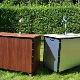 Dwa mobilne bary z kranami do piwa, jeden z drewnianym wykończeniem, drugi srebrny, stoją na trawie przed żywopłotem.