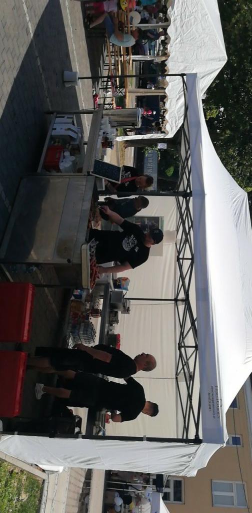 Przygotowywanie jedzenia pod białym namiotem, widoczni pracownicy w czarnych koszulkach i czapkach, serwujący dania z podgrzewacza, w tle stoliki z gośćmi.