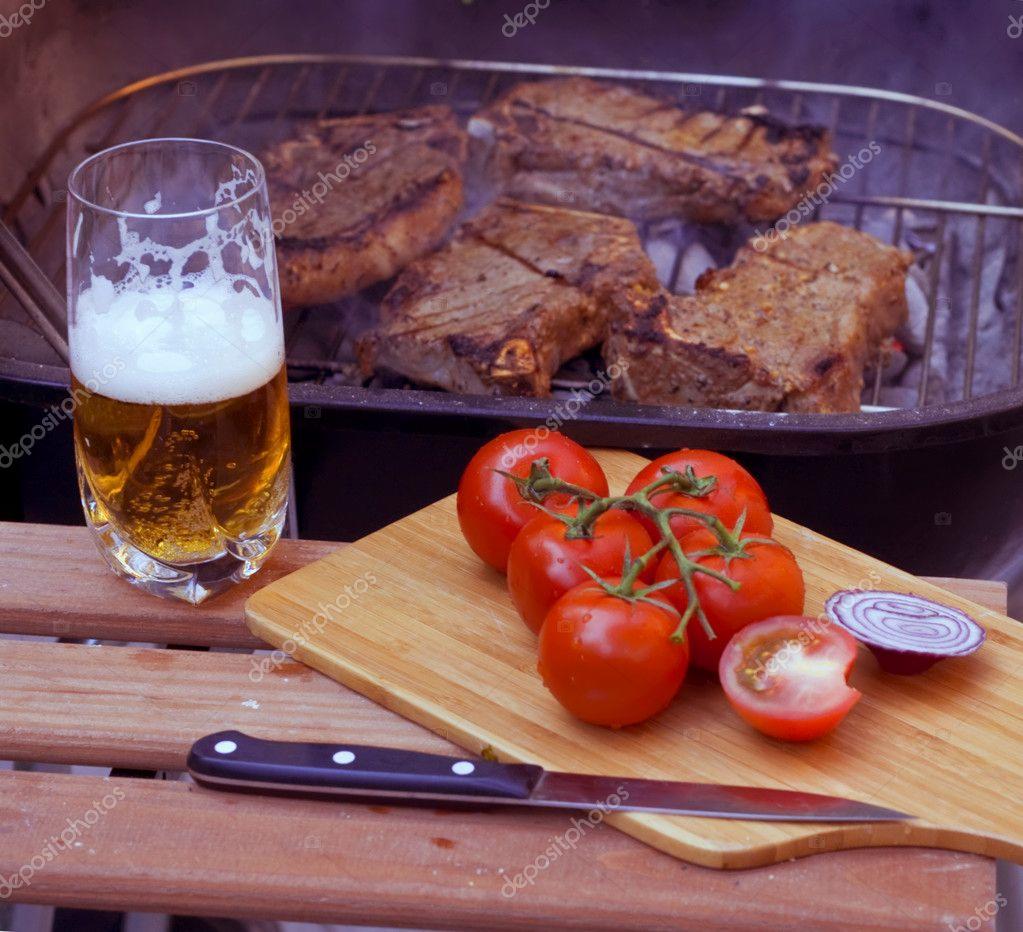 Grillowane steki wołowe na ruszcie, obok szklanka jasnego piwa, pomidory na gałązce i cebula na desce do krojenia, nóż z czarną rękojeścią na drewnianym stole.