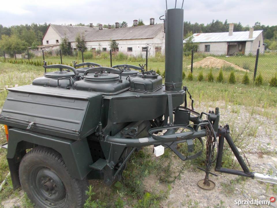 Mobilna kuchnia polowa w kolorze khaki z czterema pojemnikami na potrawy z pokrywami, z kominem i przyczepką, ustawiona na zewnątrz w plenerze.