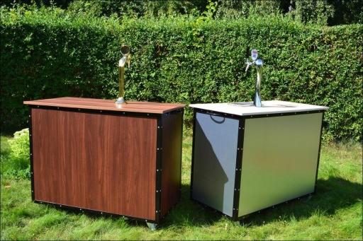 Dwa mobilne bary z kranami do piwa, jeden z drewnianym wykończeniem, drugi srebrny, stoją na trawie przed żywopłotem.