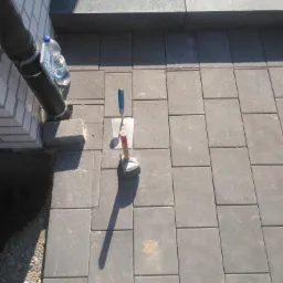 Układanie szarej kostki brukowej na zewnątrz, widoczne narzędzia: kielnia i gumowy młotek, obok rura spustowa i butelka wody, w tle schody z kostki.