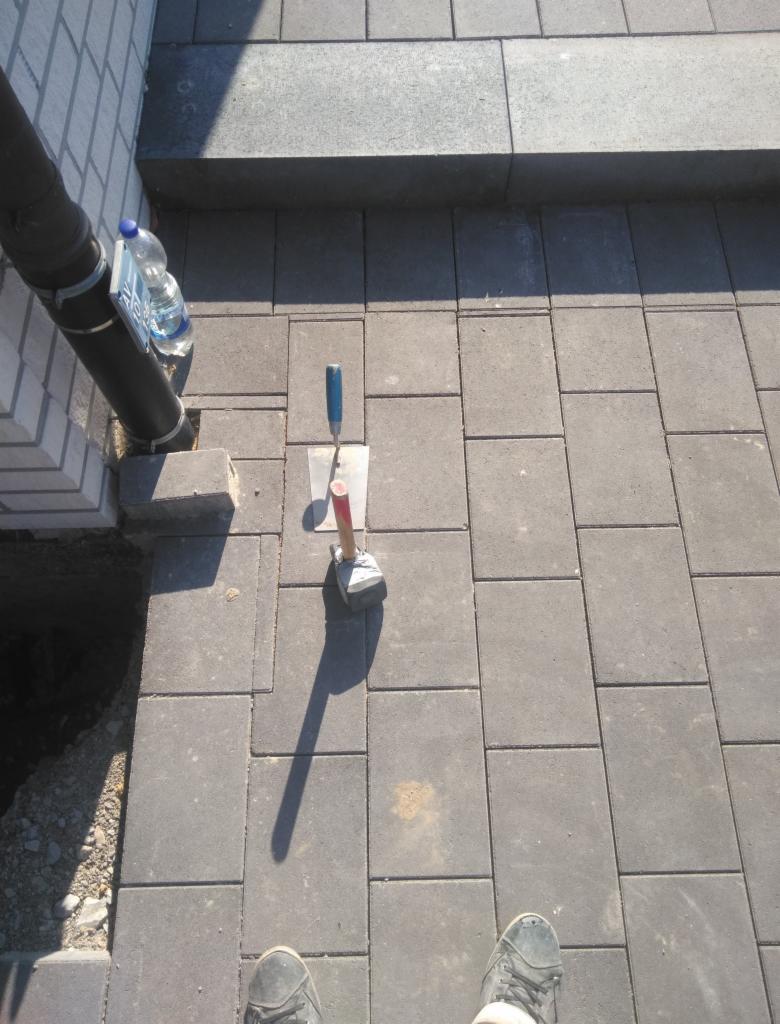 Układanie szarej kostki brukowej na zewnątrz, widoczne narzędzia: kielnia i gumowy młotek, obok rura spustowa i butelka wody, w tle schody z kostki.