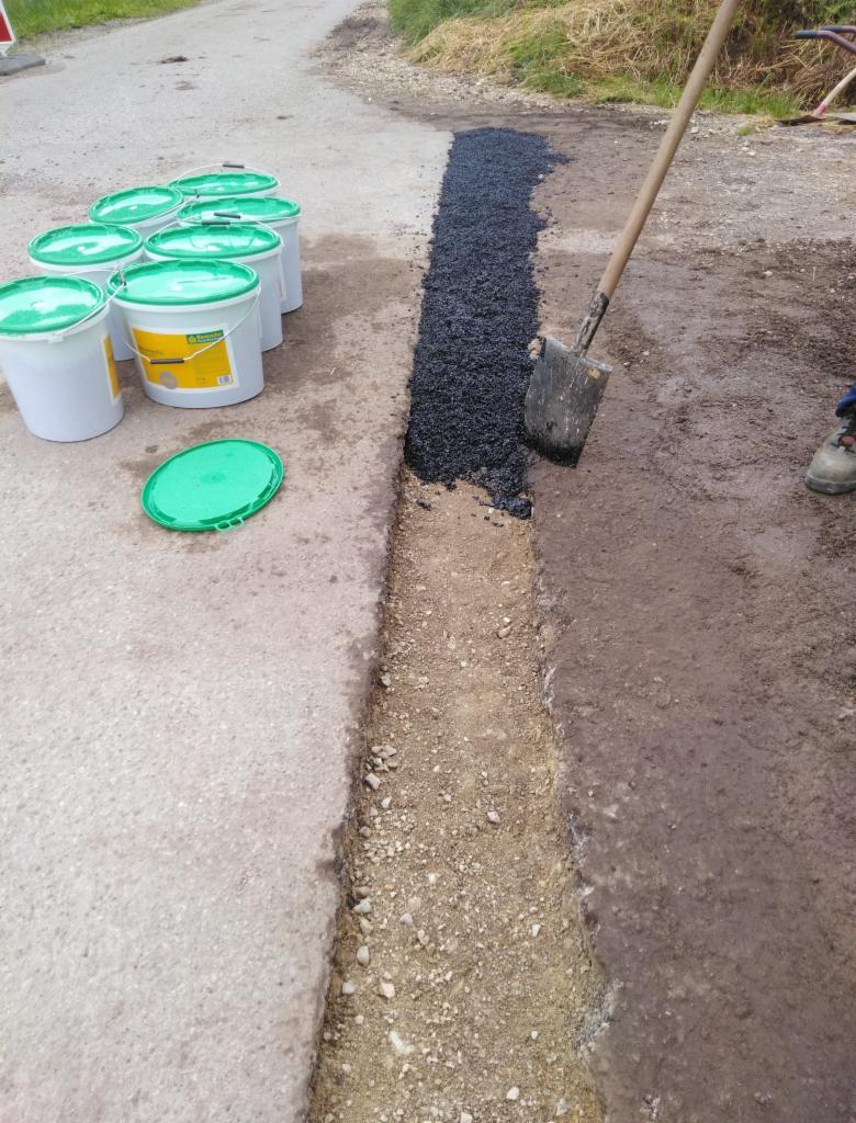 Wykopany rów wzdłuż asfaltowej drogi, wypełniany czarnym asfaltem z pomocą szpadla, obok ustawione białe wiadra z zielonymi pokrywkami.