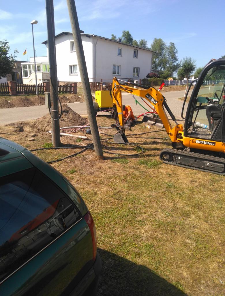 Żółta mini koparka przy słupach telegraficznych, robotnik w pomarańczowej koszulce w wykopie, widoczny agregat prądotwórczy, zarys zielonego samochodu w lewym dolnym rogu.