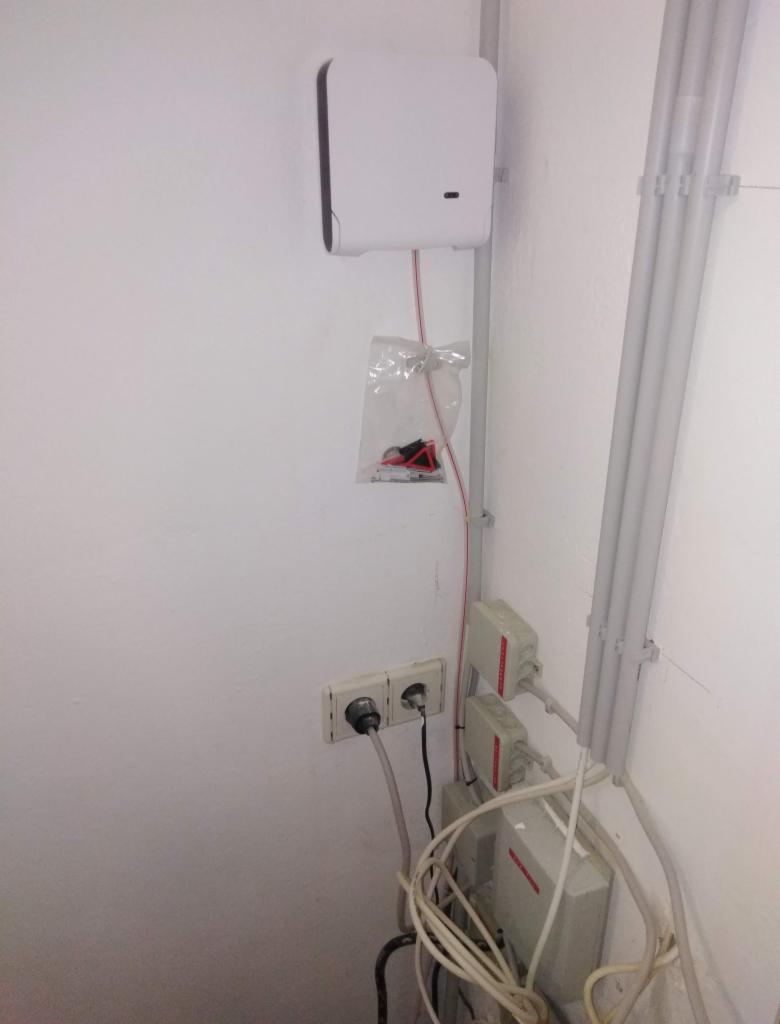 Biała skrzynka telekomunikacyjna zamontowana na ścianie, z widocznymi kablami i gniazdkami elektrycznymi w dolnej części kadru. Dodatkowe akcesoria montażowe w przezroczystej torebce wiszą...