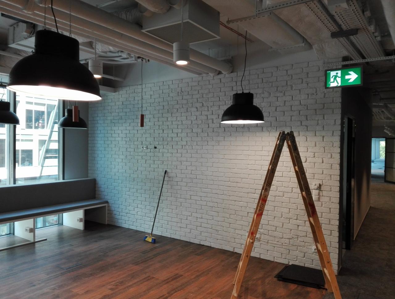 Biała ceglana ściana w biurze w Warszawie, oświetlona wiszącymi lampami, z drewnianą drabiną i miotłą opartą o ścianę, sugerująca prace wykończeniowe.