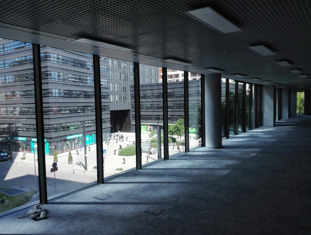 Wnętrze biurowe z dużymi oknami, widok na budynki biurowe z zewnątrz, szara wykładzina dywanowa, kolumny podtrzymujące sufit, kratownica sufitowa z oświetleniem.