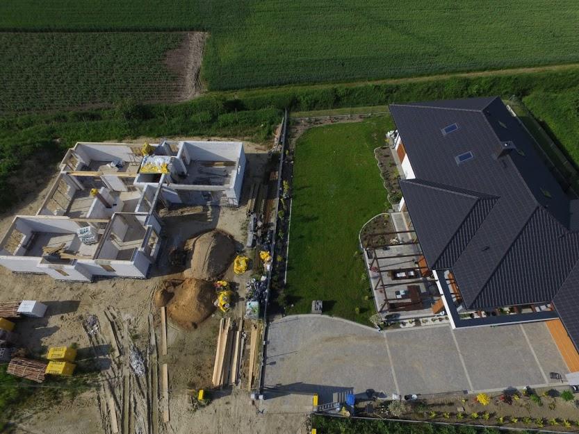 Widok z lotu ptaka na budowę domu jednorodzinnego obok już istniejącego, wykończonego domu z czarnym dachem, widoczny trawnik i przygotowany teren pod ogród.