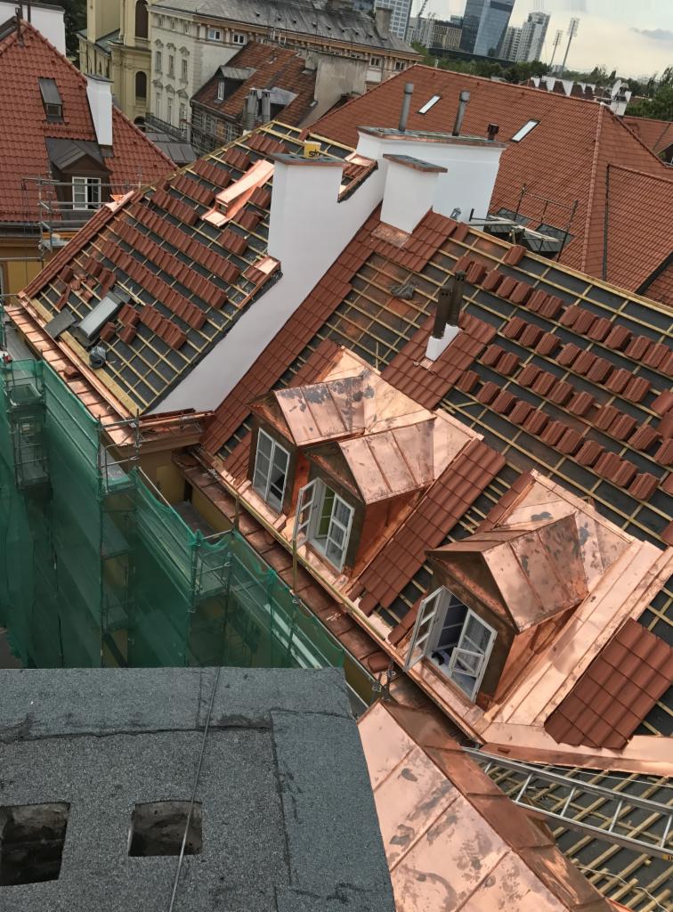 Wysoki kąt widzenia na dach budynku w trakcie renowacji, z częściowo ułożoną czerwoną dachówką i miedzianymi lukarnami, widoczne rusztowania z zieloną siatką zabezpieczającą.