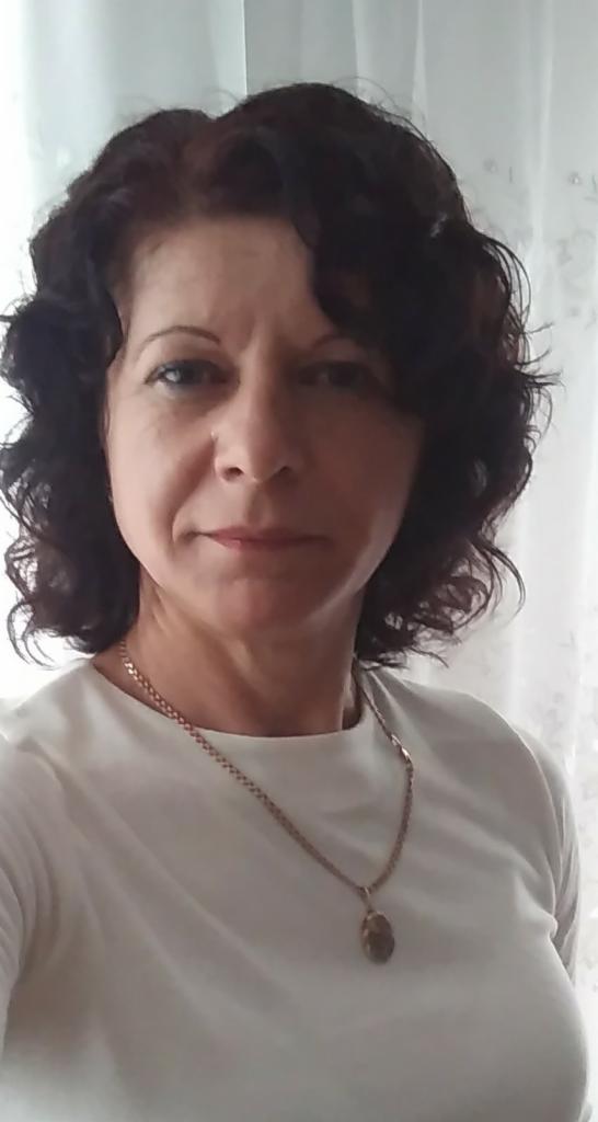 Portret kobiety o ciemnych, kręconych włosach, w białej bluzce i złotej biżuterii. Uśmiech i delikatne zmarszczki wokół oczu. Naturalne światło.