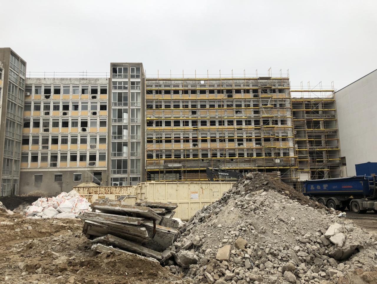 Widok na budynek w trakcie rozbiórki, z rusztowaniami, stertą gruzu, workami z odpadami i ciężarówką w tle.