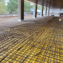 Zbrojenie płyty fundamentowej z widocznymi prętami zbrojeniowymi ułożonymi na żółtej folii izolacyjnej, wsparte na kolumnach z szarego betonu.