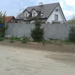 Nowoczesny dom z szarą dachówką, białymi ścianami i betonowym ogrodzeniem, widoczny z ulicy Rolna