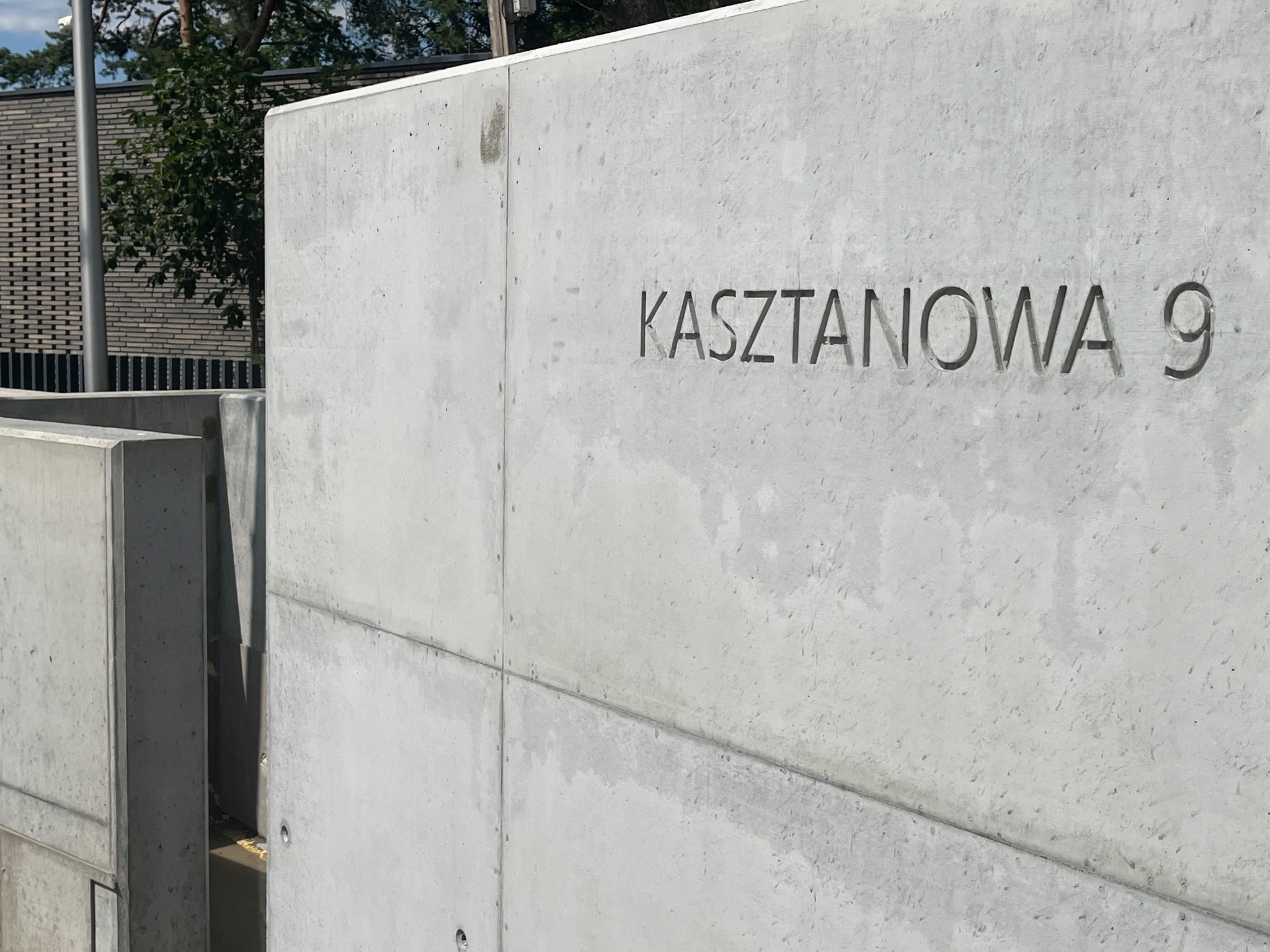 Betonowa ściana z wygrawerowanym adresem 'Kasztanowa 9', widoczna faktura betonu i łączenia płyt, w tle fragment budynku z cegły.