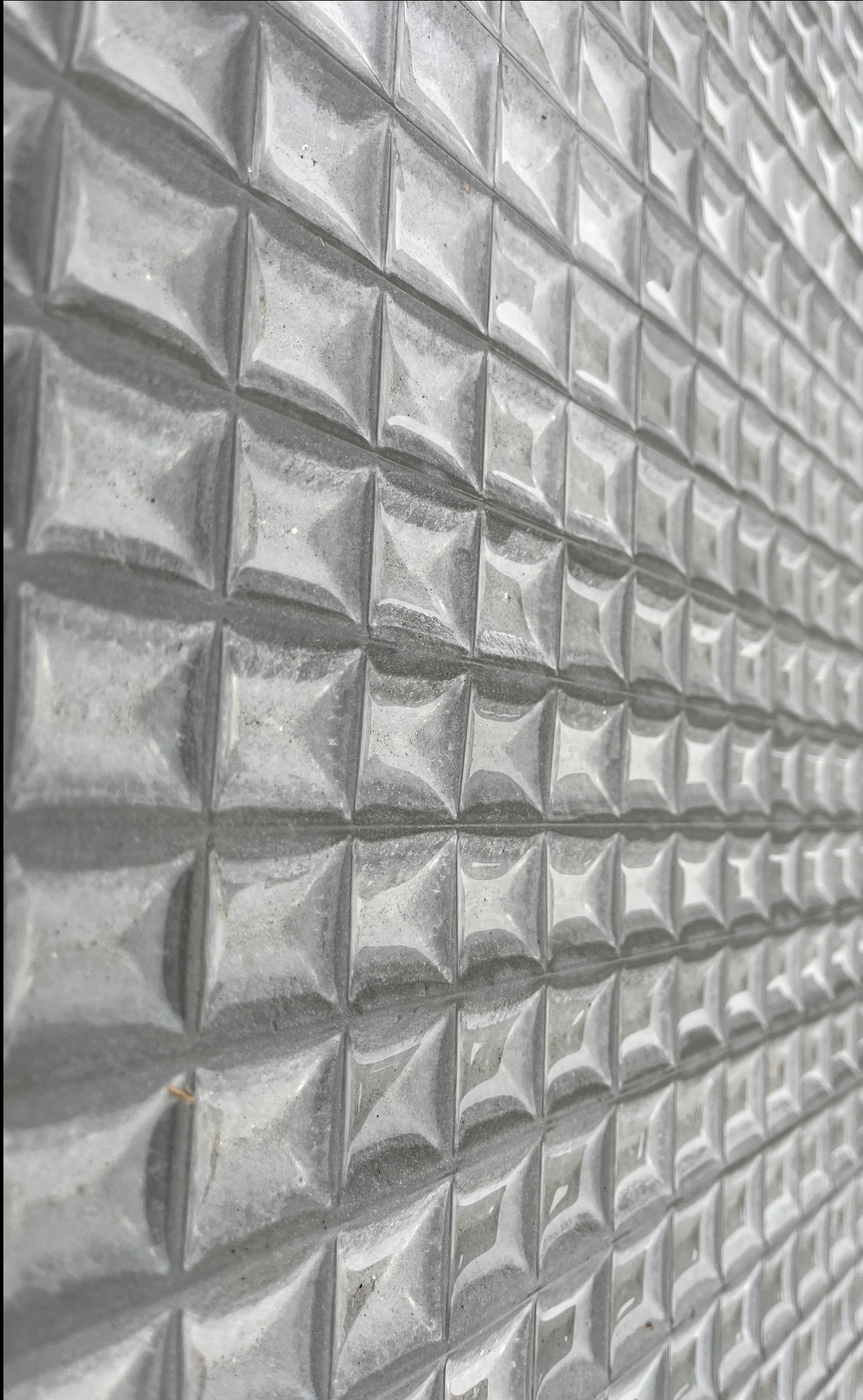 Szara betonowa ściana z regularnym wzorem kwadratowych wypukłości, ułożonych pod kątem, z widocznymi śladami po szalunku.