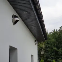 Dwa chromowane wyloty wentylacyjne na białej elewacji budynku, widok z dołu, obok zaparkowany biały bus.