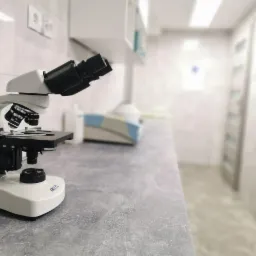 Mikroskop laboratoryjny Delta na blacie w sterylnym pomieszczeniu, w tle widoczny sprzęt medyczny i przeszklone drzwi.