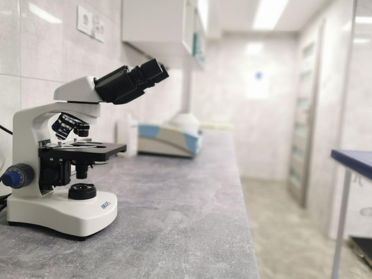 Mikroskop laboratoryjny Delta na blacie w sterylnym pomieszczeniu, w tle widoczny sprzęt medyczny i przeszklone drzwi.