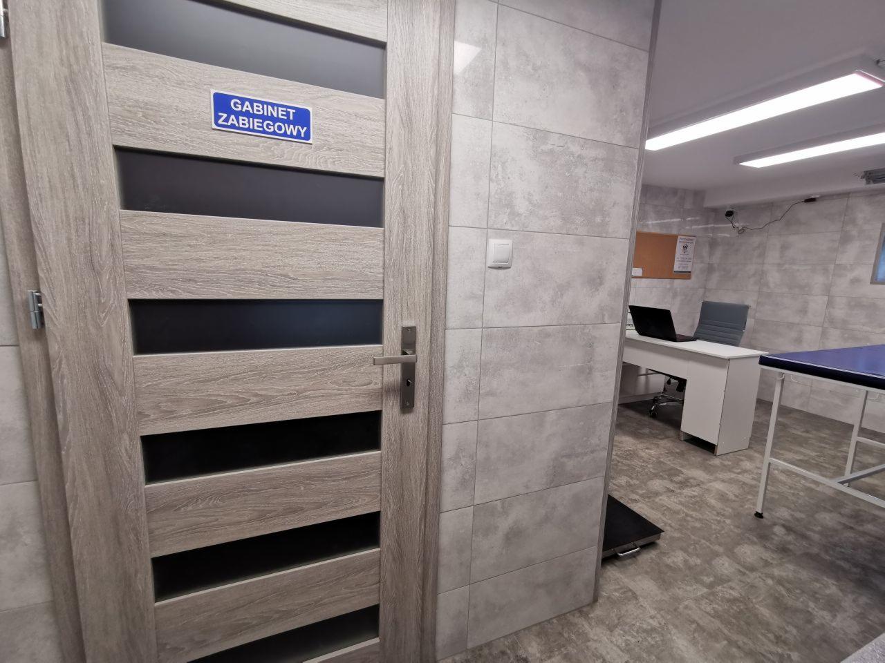 Wejście do gabinetu zabiegowego z drewnopodobnymi drzwiami i niebieską tabliczką informacyjną, widoczny fragment wnętrza z biurkiem, laptopem i stołem zabiegowym.