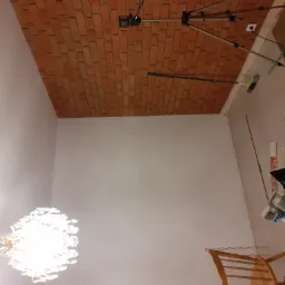 Ceglana okładzina sufitowa w trakcie montażu, widoczny laserowy niwelator na statywie, narzędzia i materiały budowlane na podłodze, kryształowy żyrandol w dolnej części kadru.