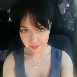 Portret kobiety z krótkimi czarnymi włosami i srebrnym naszyjnikiem z literą K, siedzącej w samochodzie z ciemną tapicerką.