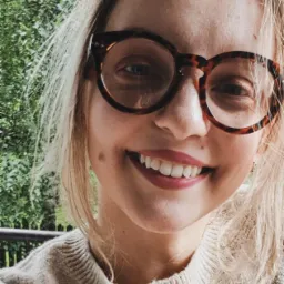 Portret uśmiechniętej blondynki w okularach o wzorze panterki na tle zieleni. Ujęcie z bliska, z widocznymi detalami skóry i włosów.