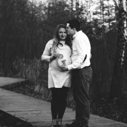 Czarno-białe zdjęcie przyszłej mamy trzymającej literę 'G' na brzuchu, partner całuje ją w czoło, stoją na drewnianej ścieżce w parku.