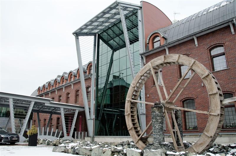 Odrestaurowany budynek z czerwonej cegły z nowoczesną szklaną dobudówką i dekoracyjnym kołem młyńskim na tle zimowego krajobrazu.