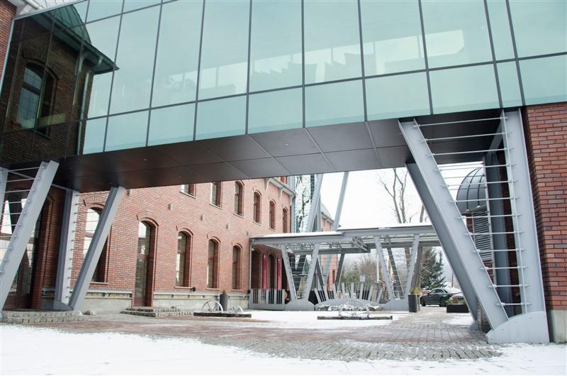 Szklany łącznik między budynkami z czerwonej cegły, wsparty na stalowych podporach, z elementami dekoracyjnymi na dziedzińcu pokrytym śniegiem.