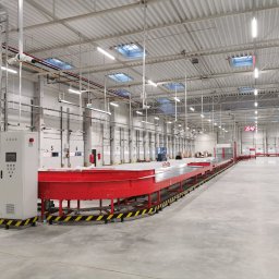 EXEVER SPÓŁKA Z OGRANICZONĄ ODPOWIEDZIALNOŚCIĄ - Wnętrze hali magazynowej z systemem przenośników taśmowych w kolorze czerwonym, panelem sterowania i bramami załadunkowymi. Oświetlenie sufitowe i betonowa posadzka.