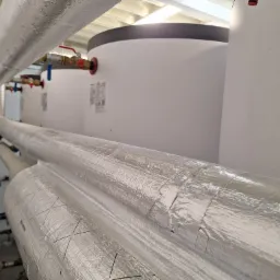 Instalacja grzewcza: rury z izolacją termiczną i zbiorniki akumulacyjne w kotłowni. Widoczne zawory i połączenia. Ujęcie z dołu, podkreślające skalę instalacji.