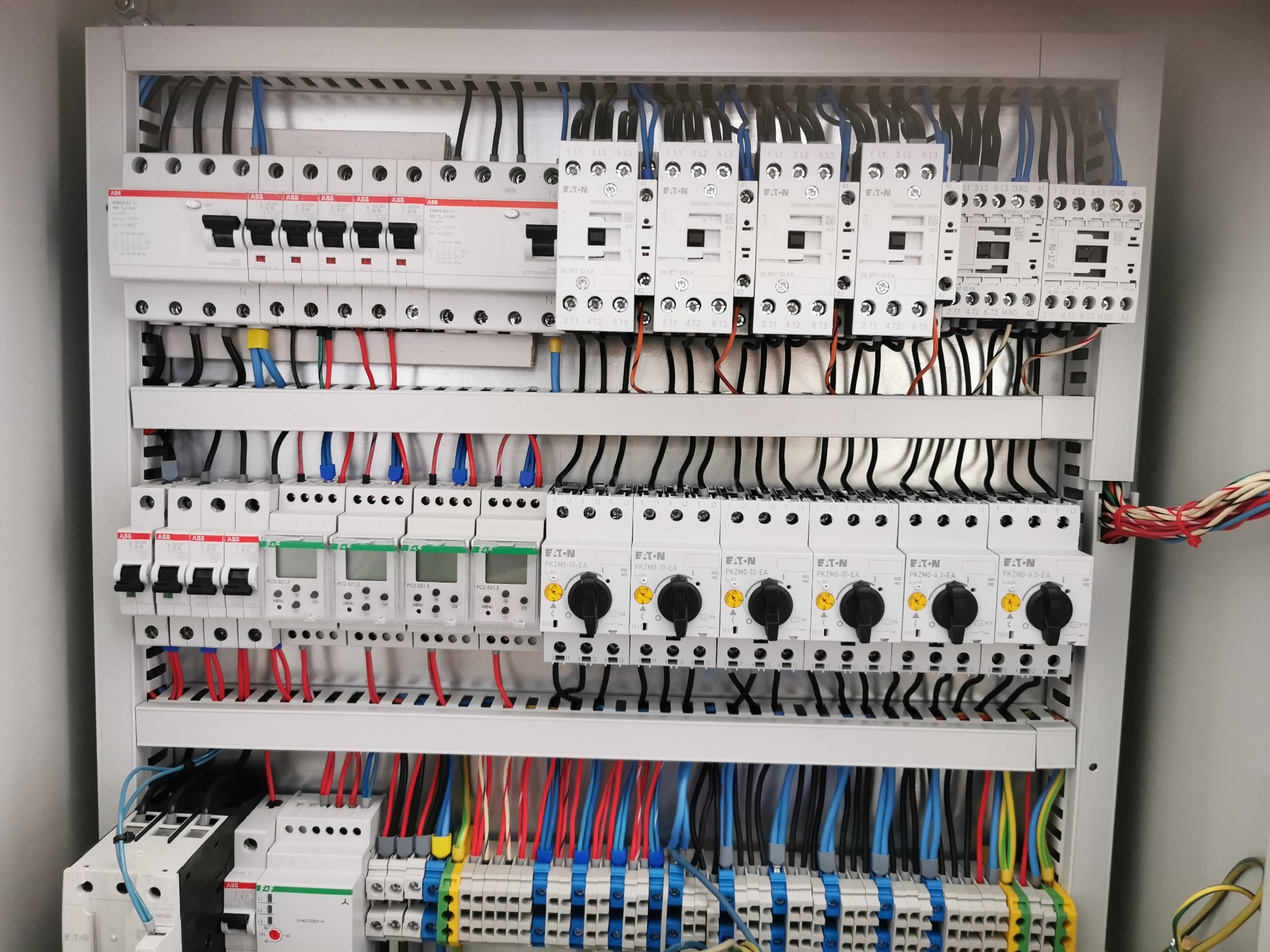 Rozdzielnia elektryczna z bezpiecznikami, stycznikami i przewodami w kolorach: czarnym, czerwonym i niebieskim. Widoczne elementy marki ABB i Eaton.