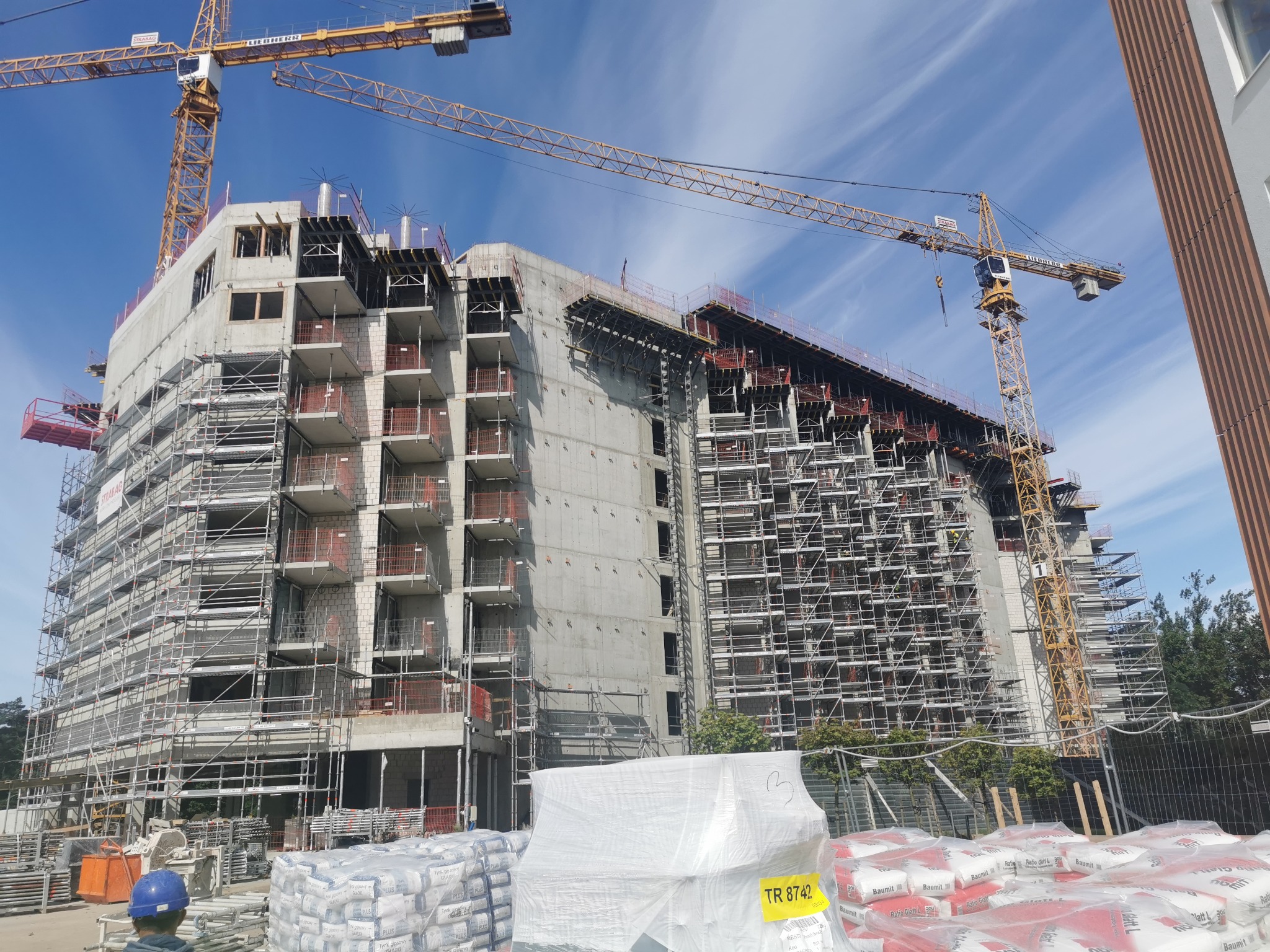 Budowa wysokiego budynku mieszkalnego w trakcie realizacji, z rusztowaniami i dźwigami na tle błękitnego nieba z chmurami. Widoczne worki z materiałami budowlanymi.