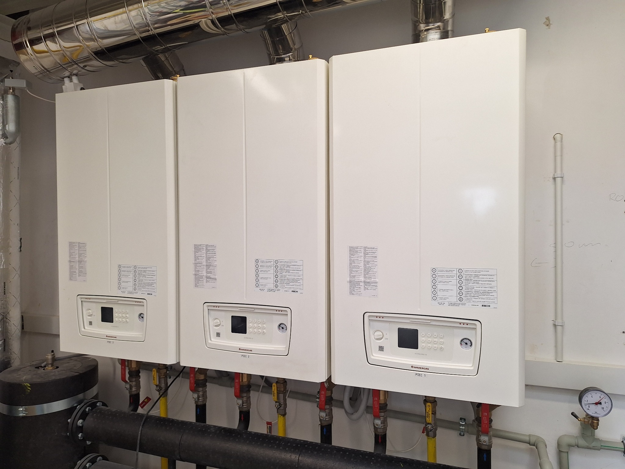 Trzy białe, wiszące piece gazowe Immergas, każdy z panelem sterowania i podłączonymi rurami. Widoczne rury instalacyjne i elementy kotłowni.