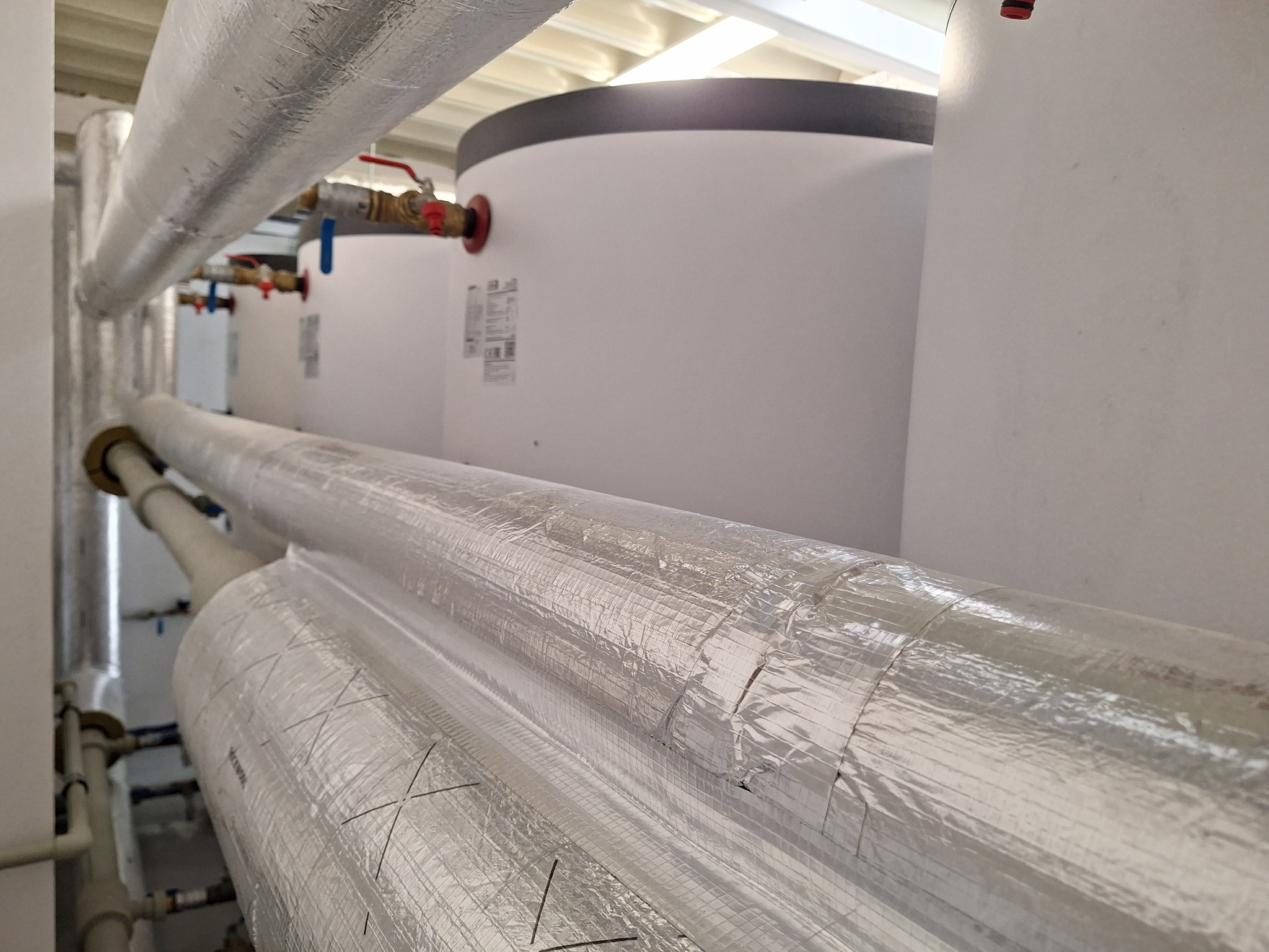 Instalacja grzewcza: rury z izolacją termiczną i zbiorniki akumulacyjne w kotłowni. Widoczne zawory i połączenia. Ujęcie z dołu, podkreślające skalę instalacji.