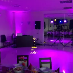 Sala bankietowa oświetlona fioletowym światłem, z widocznym sprzętem nagłośnieniowym, stołami nakrytymi do uroczystości i odbiciem w lustrze dalszej części sali.